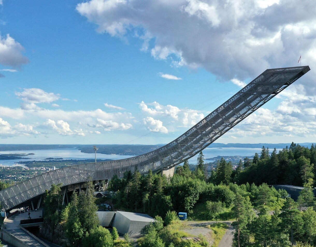 Aerial view of Holmenkollen Ski Jump in Oslo, Norway, surrounded by green forest with Oslofjord and cityscape in the distance under a bright blue sky.