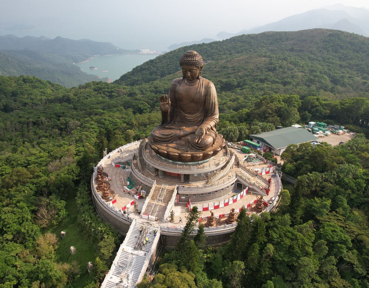 Aerial view of the Tian Tan Buddha statue on Lantau Island, Hong Kong, showing the large bronze Buddha seated atop a circular terrace surrounded by dense green forest and distant mountain and ocean views.