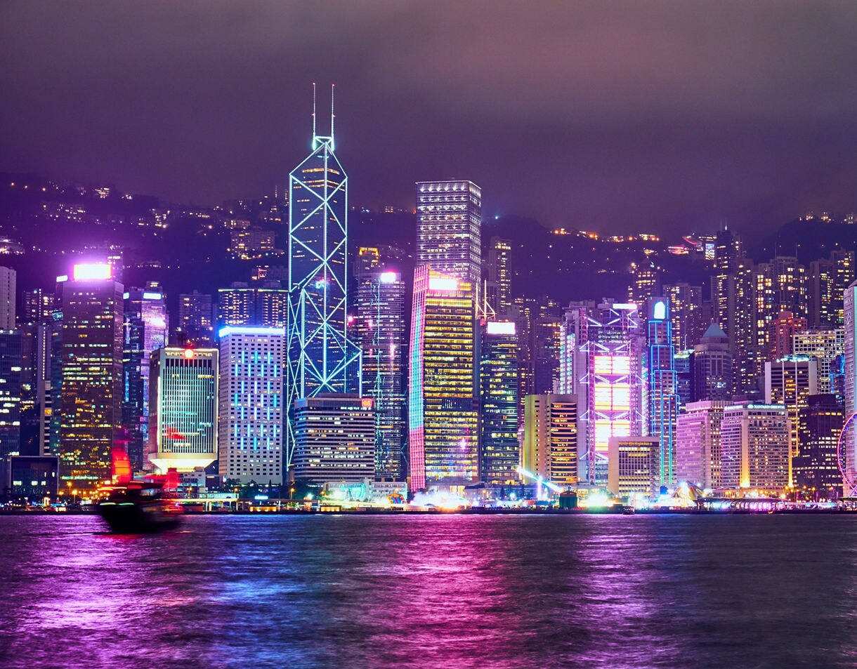 Night view of Hong Kong’s skyline with brightly lit neon skyscrapers reflected in Victoria Harbour, including a ferris wheel and a blurred boat moving across the foreground.