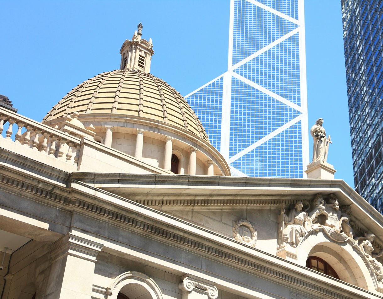 Close-up view of Hong Kong’s historic Legislative Council Building with its domed roof and stone statues, framed against tall modern skyscrapers and a bright blue sky.