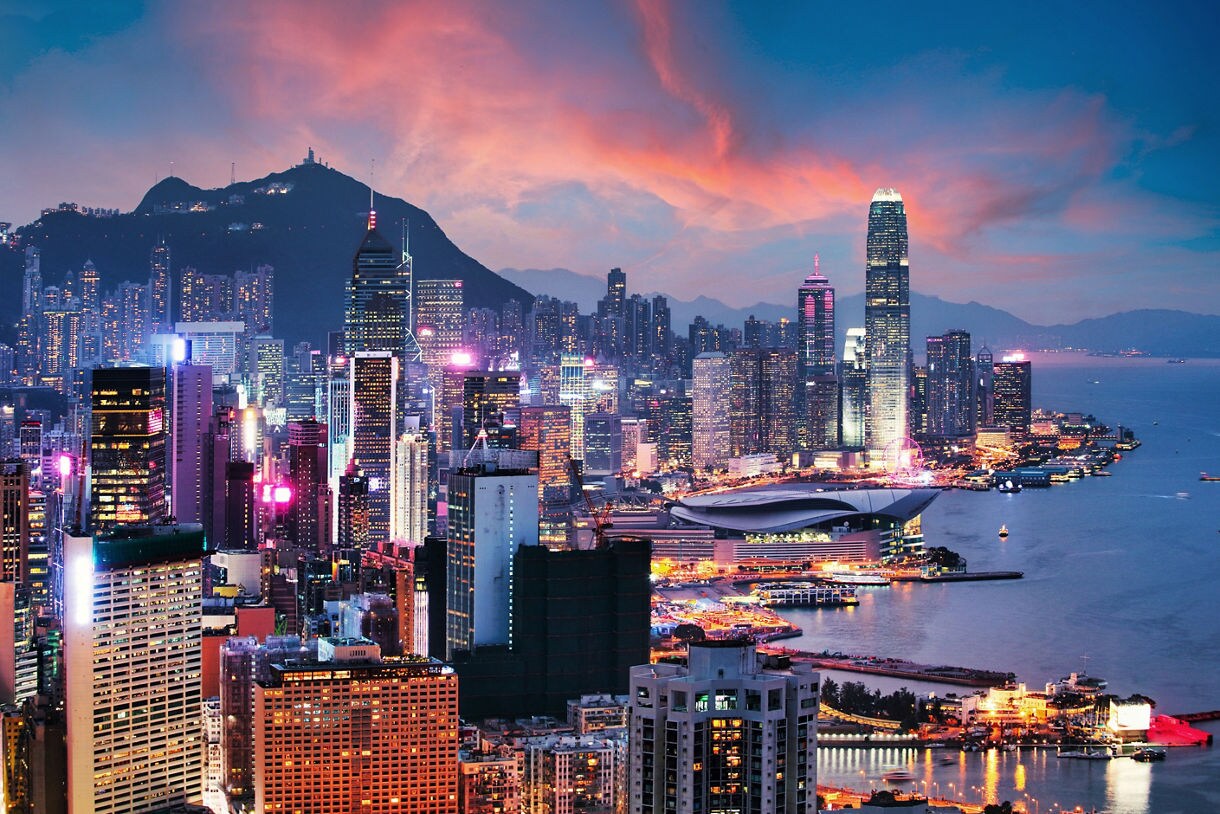 Panoramic view of Hong Kong’s skyline at sunset, featuring illuminated skyscrapers, Victoria Harbour, and pastel clouds glowing above the city.