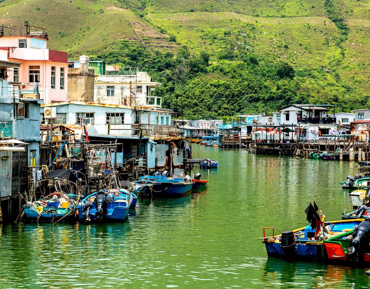 View of a traditional fishing village on Lantau Island, Hong Kong, with stilt houses along green water and brightly colored small boats moored in the canal, set against lush hillside scenery.