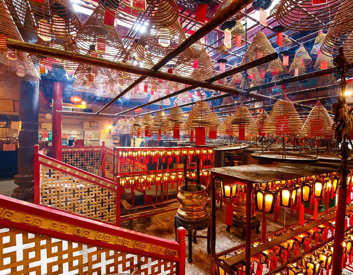 Interior of Man Mo Temple in Hong Kong filled with hanging spiral incense coils, red prayer tags, glowing lanterns and ornate red railings, creating a warm and smoky atmosphere.