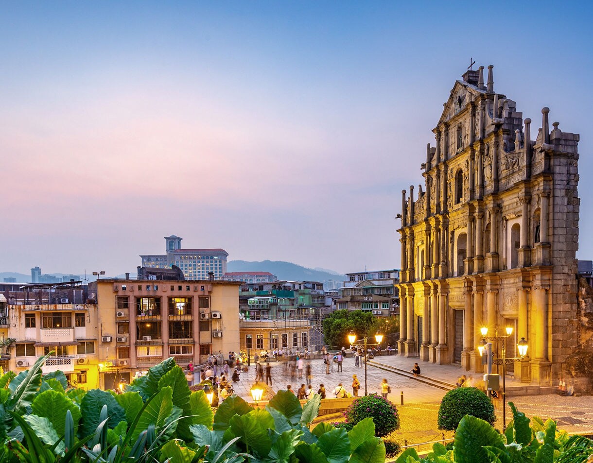 View of the Ruins of St. Paul’s in Macau at sunset, with the illuminated stone façade overlooking a busy square and surrounding city buildings framed by lush green plants.