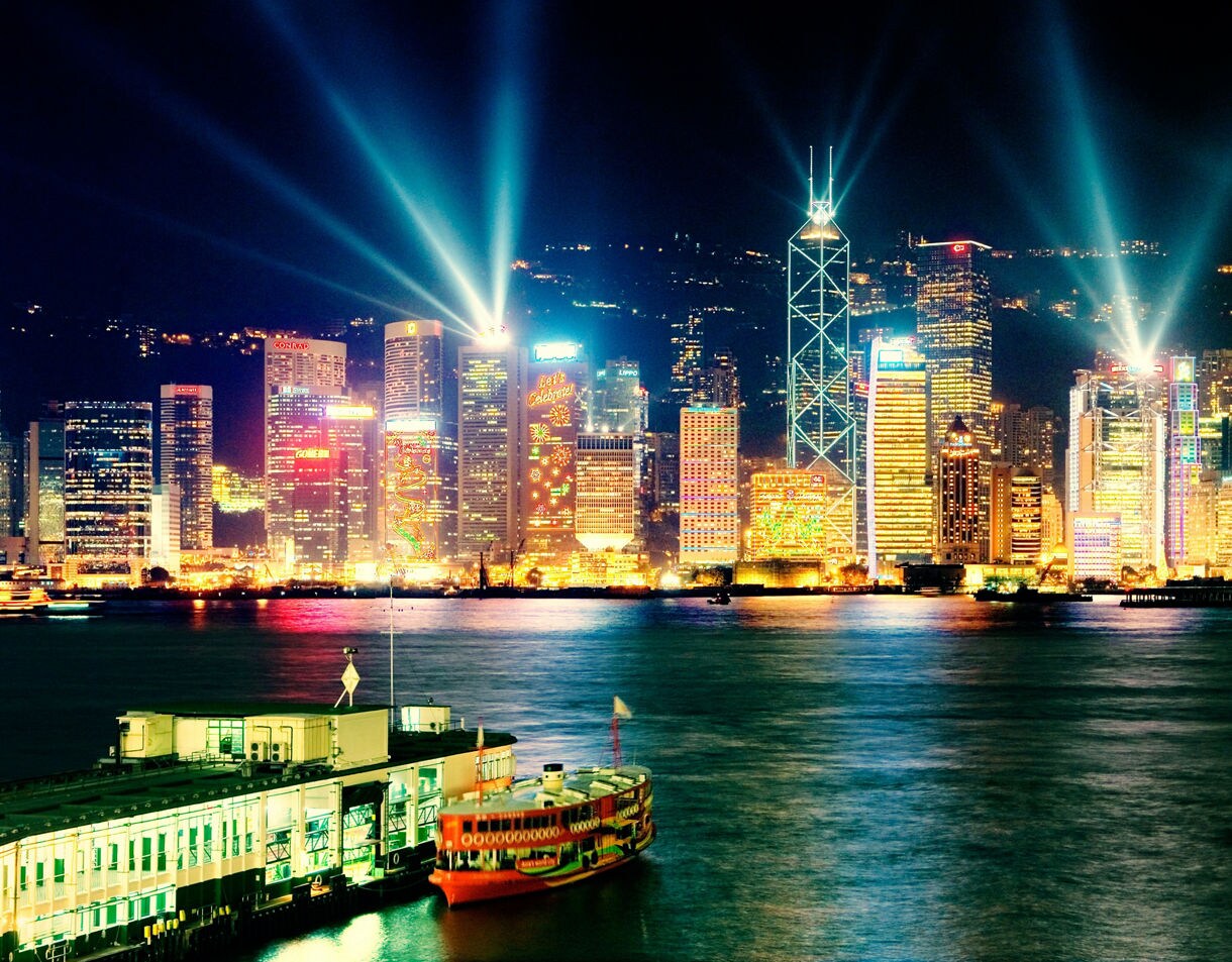 Night view of Hong Kong’s illuminated skyline with colorful laser lights shining from tall buildings and a brightly painted Star Ferry crossing Victoria Harbour in the foreground.