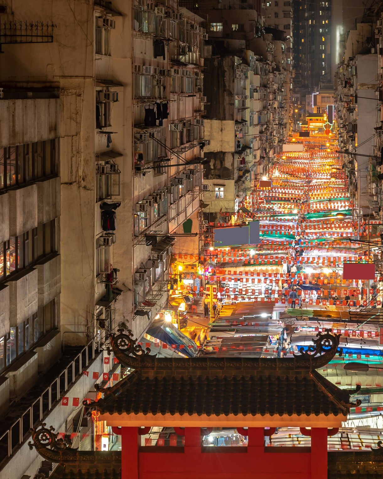 A nighttime view of Temple Street Market in Hong Kong, framed between tall apartment buildings with rows of brightly lit market stalls stretching into the distance beneath colorful awnings and lanterns.