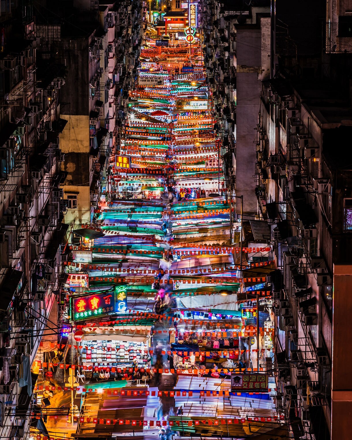 Overhead view of Hong Kong’s Temple Street Night Market at night, with rows of colorful canopy-covered stalls, neon signs and crowds moving between tightly packed buildings.