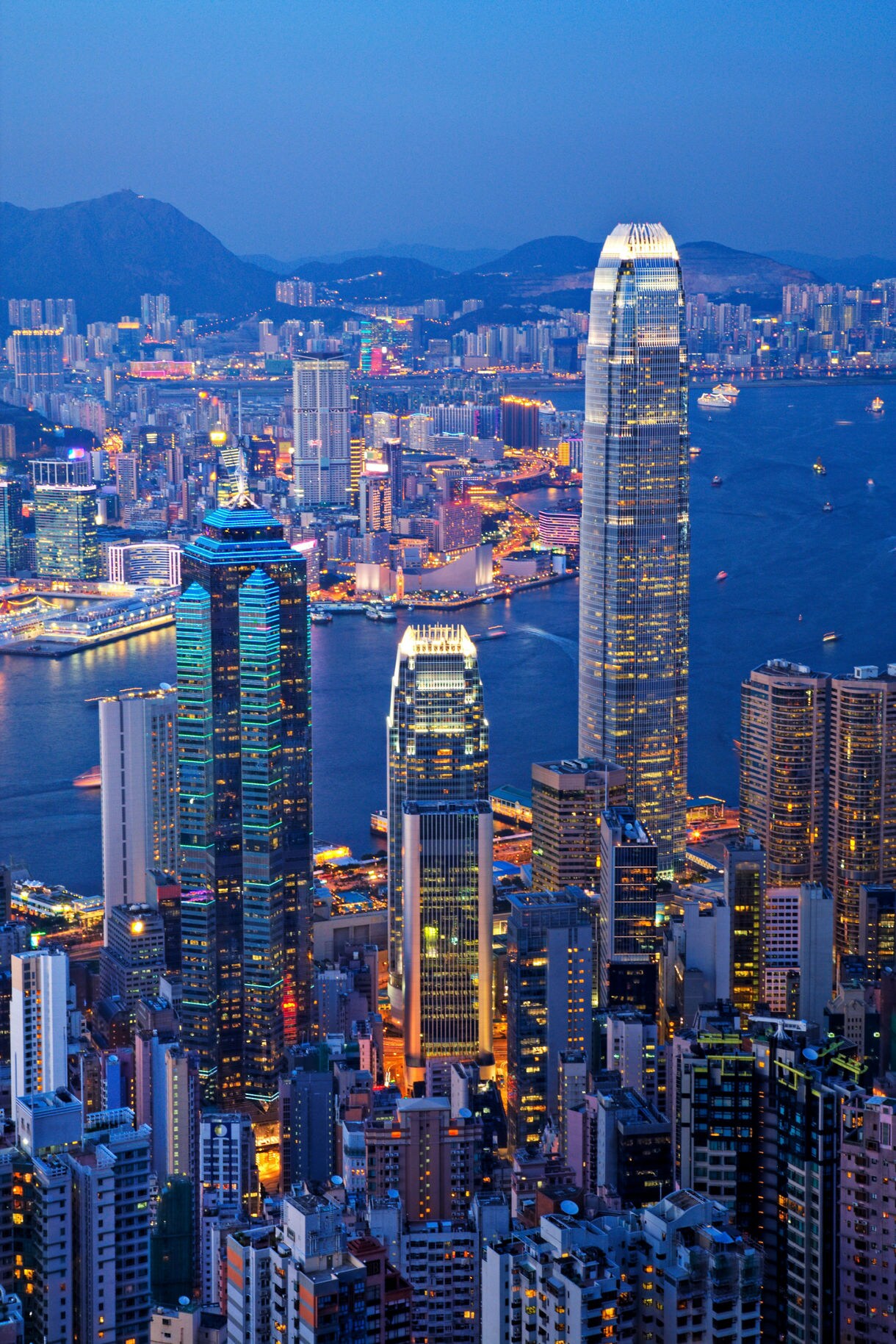 Aerial evening view of Hong Kong’s skyline with illuminated skyscrapers rising along Victoria Harbour, backed by distant mountains and city lights reflecting on the water.