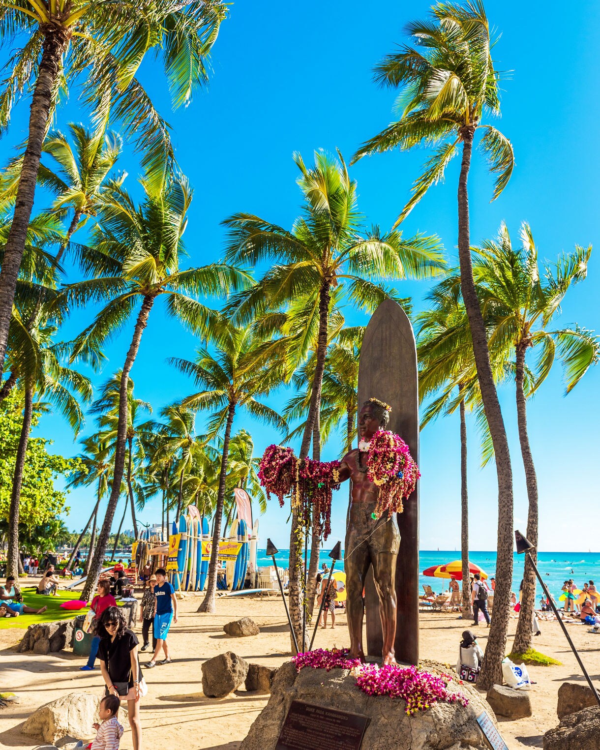 Bronze statue of Duke Kahanamoku draped in flower leis, standing before a surfboard on Waikiki Beach surrounded by palm trees.