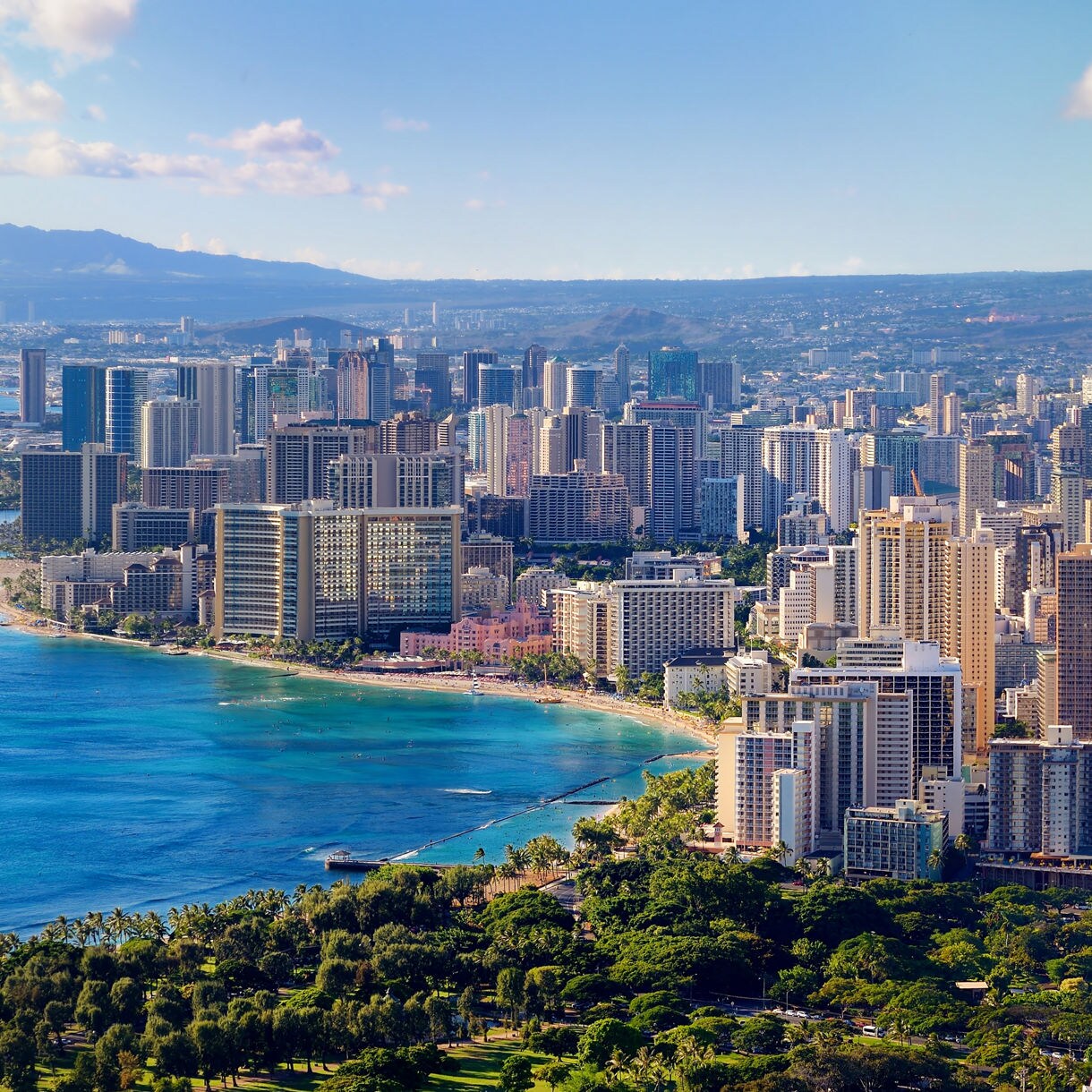 Aerial view of Honolulu with tall buildings lining Waikiki Beach, turquoise ocean on the left and lush mountains in the distance.
