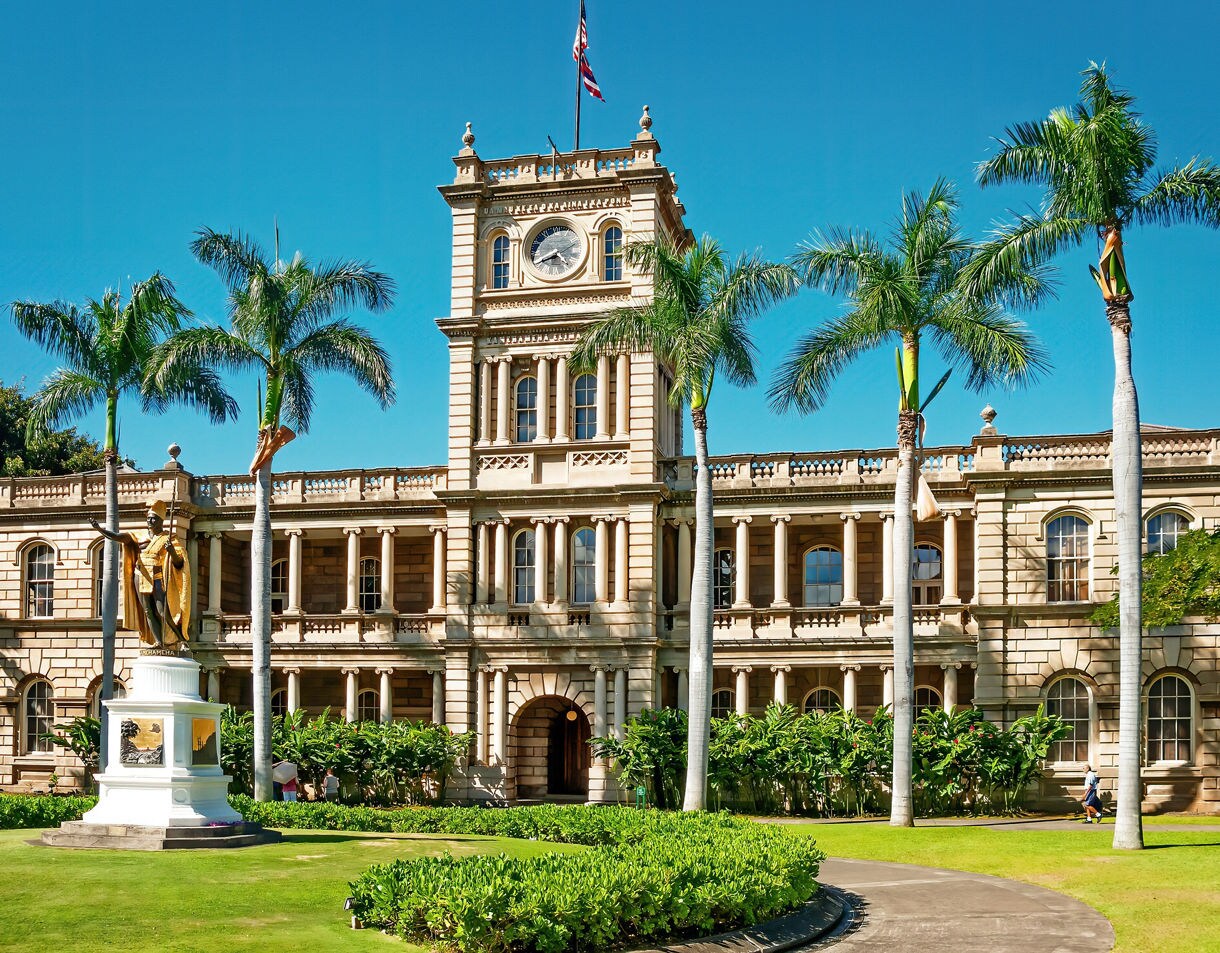 Exterior of ʻIolani Palace in Honolulu with palm trees, clock tower, and a gold King Kamehameha statue in front.