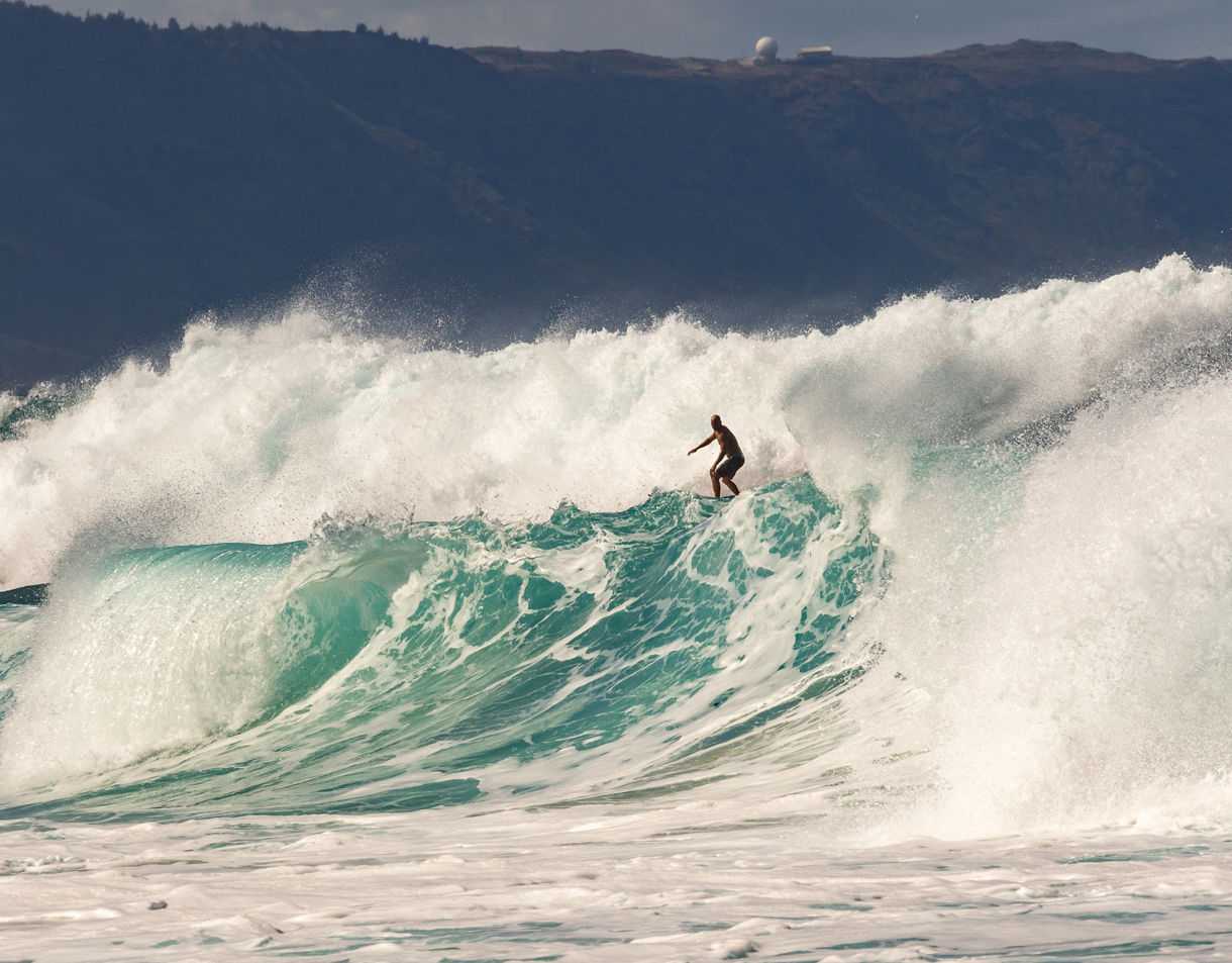 Surfer balancing on a massive turquoise wave with white spray crashing around at Oahu’s North Shore.