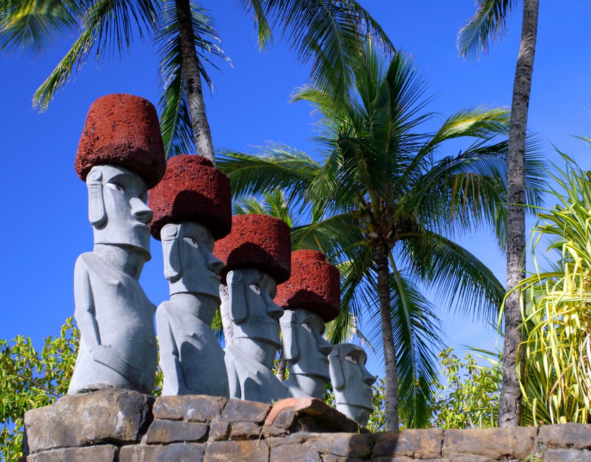 Row of Polynesian stone statues with red headpieces set against palm trees and blue sky at the Polynesian Cultural Center.