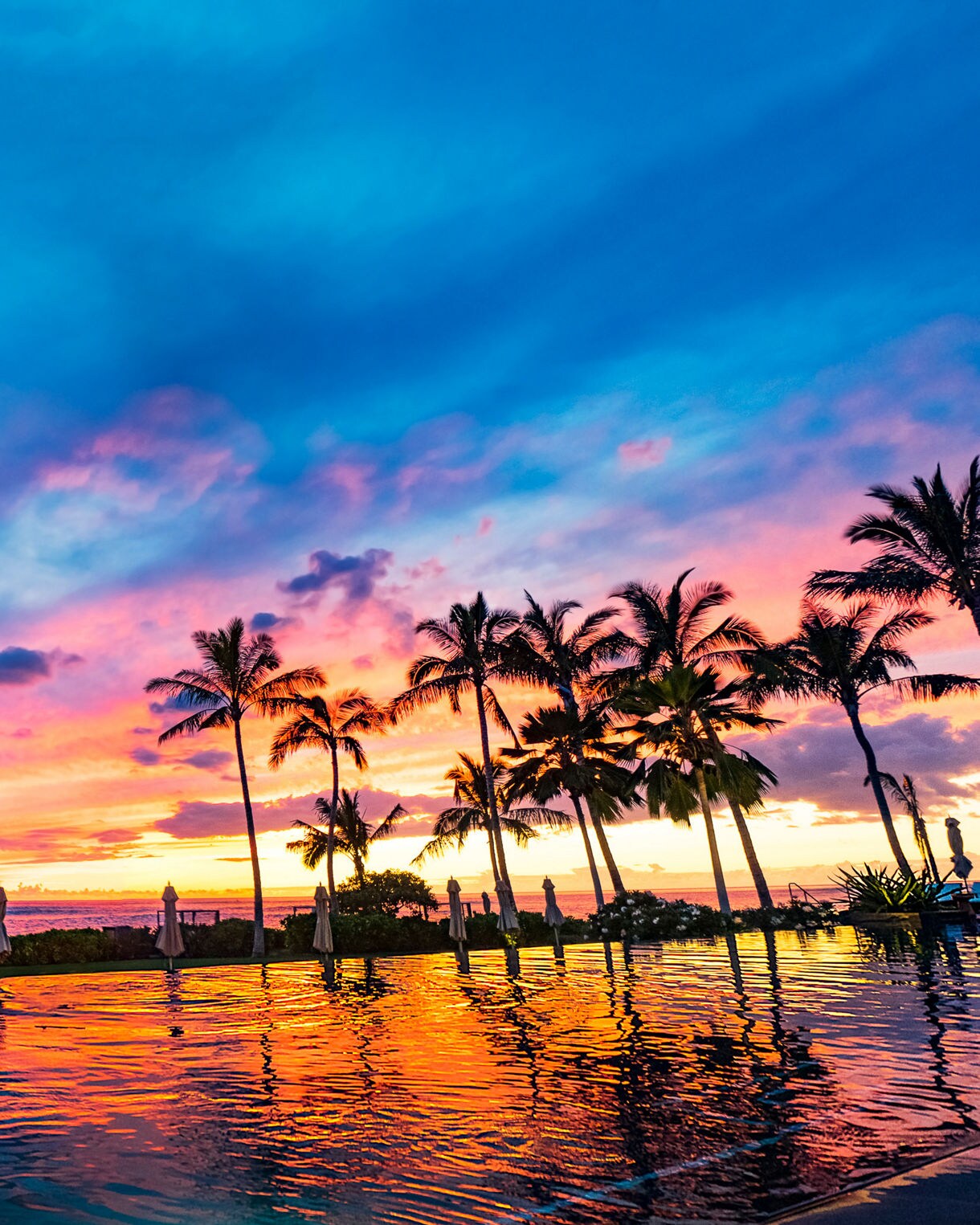 A tranquil infinity pool reflects a vivid tropical sunset with dramatic clouds and rich colors in the sky. Tall palm trees line the water’s edge, silhouetted against shades of blue, pink, orange, and purple. Lounge chairs and umbrellas suggest a luxurious resort setting by the ocean. The calm water and expansive horizon create a serene, vacation-like atmosphere.