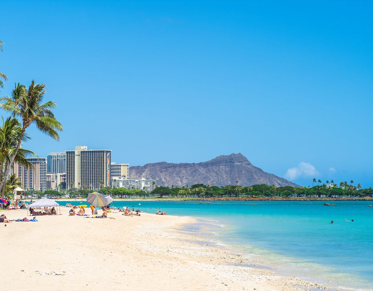 Waikiki Beach with sunbathers under umbrellas, turquoise water, and Diamond Head crater rising beyond the city skyline.