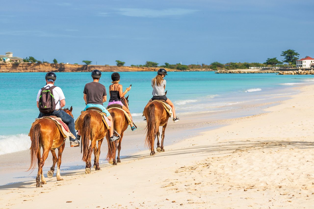 A group of people riding horses on the sandy beach of St. Maarten, with clear blue skies, turquoise water and a row of palm trees in the background.