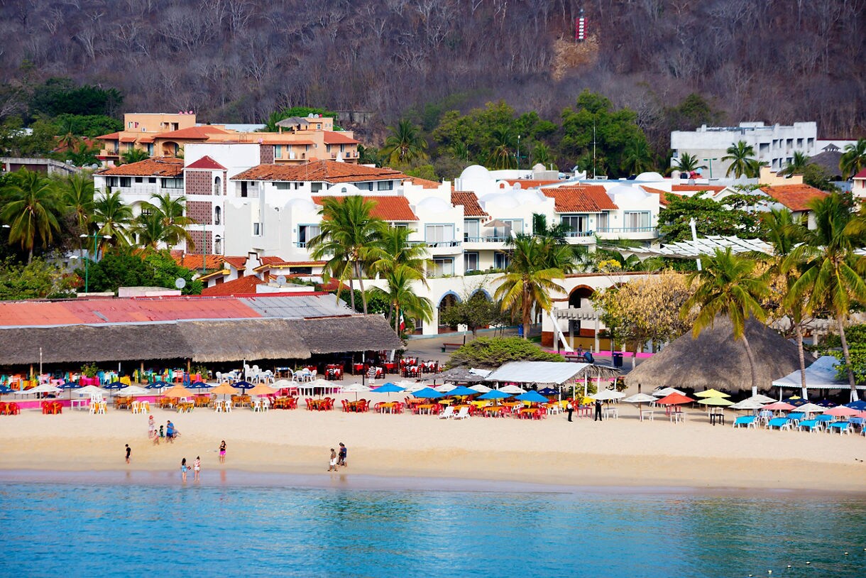 Beachfront at Huatulco Bay, Mexico with rows of vibrant umbrellas and chairs on the sand, backed by palm trees, whitewashed buildings and red-tiled roofs.