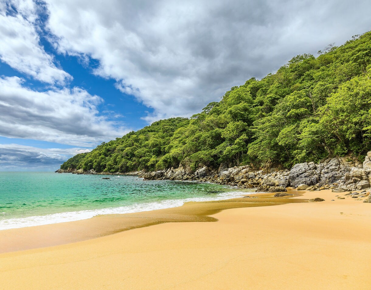 Secluded beach in Huatulco with golden sand, emerald-green water and rocky cliffs covered in dense tropical forest under a partly cloudy sky.