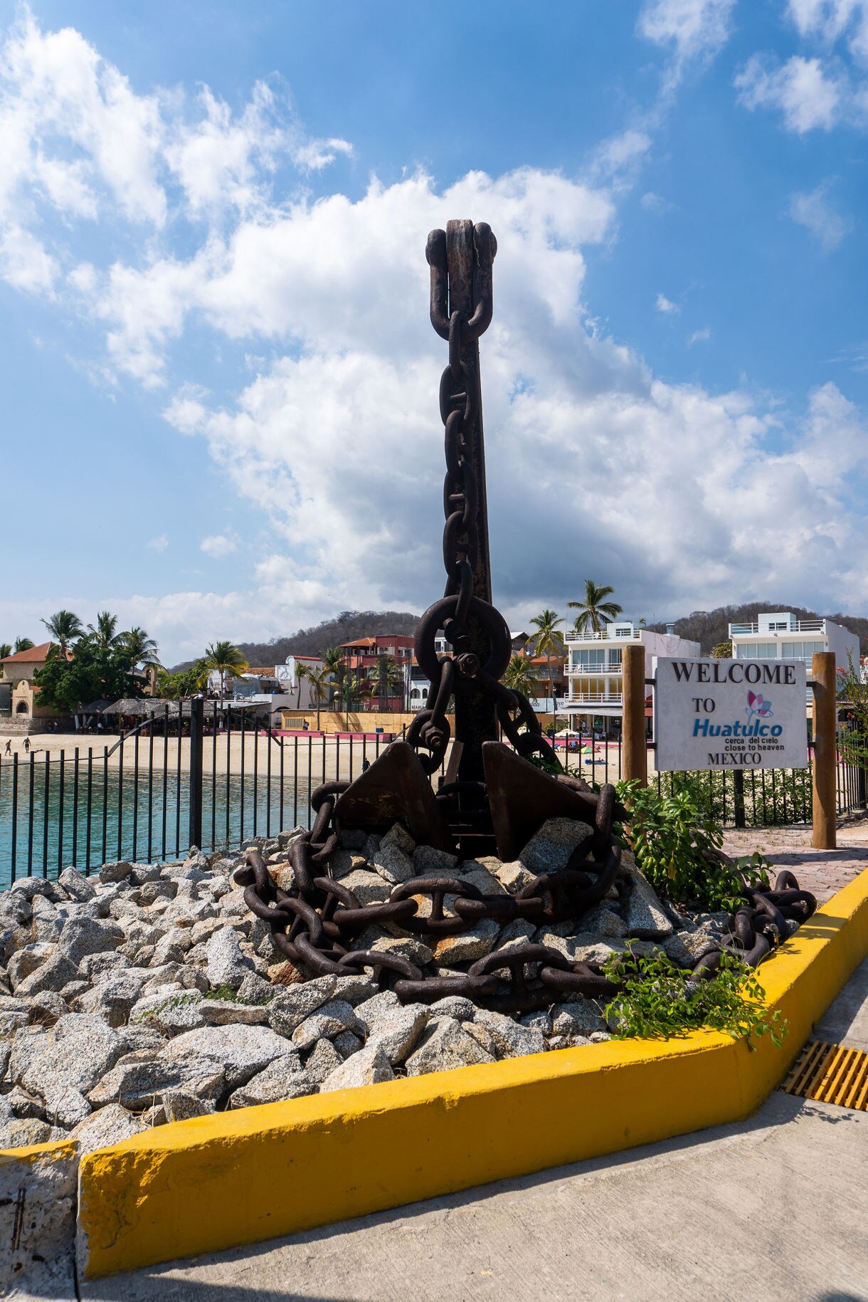 Large iron anchor monument set on rocks by the waterfront in Huatulco, Oaxaca, with a welcome sign and beachside buildings in the background.