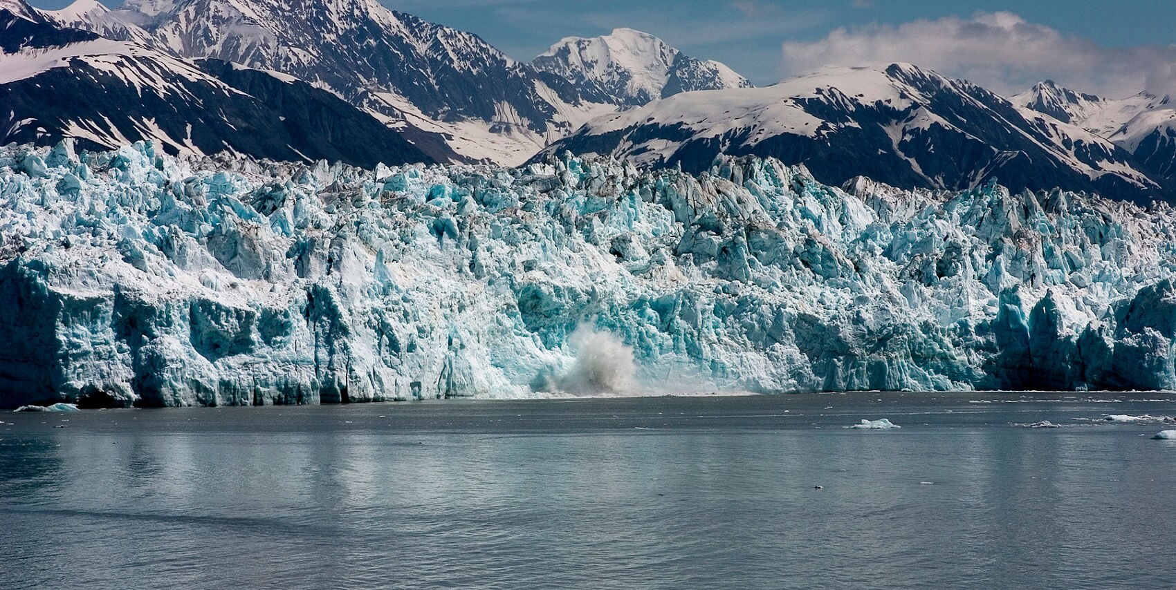 The massive turquoise blue face of Hubbard Glacier rises from dark water with a splash of ice and mist near its base, backed by snow-covered mountain peaks.