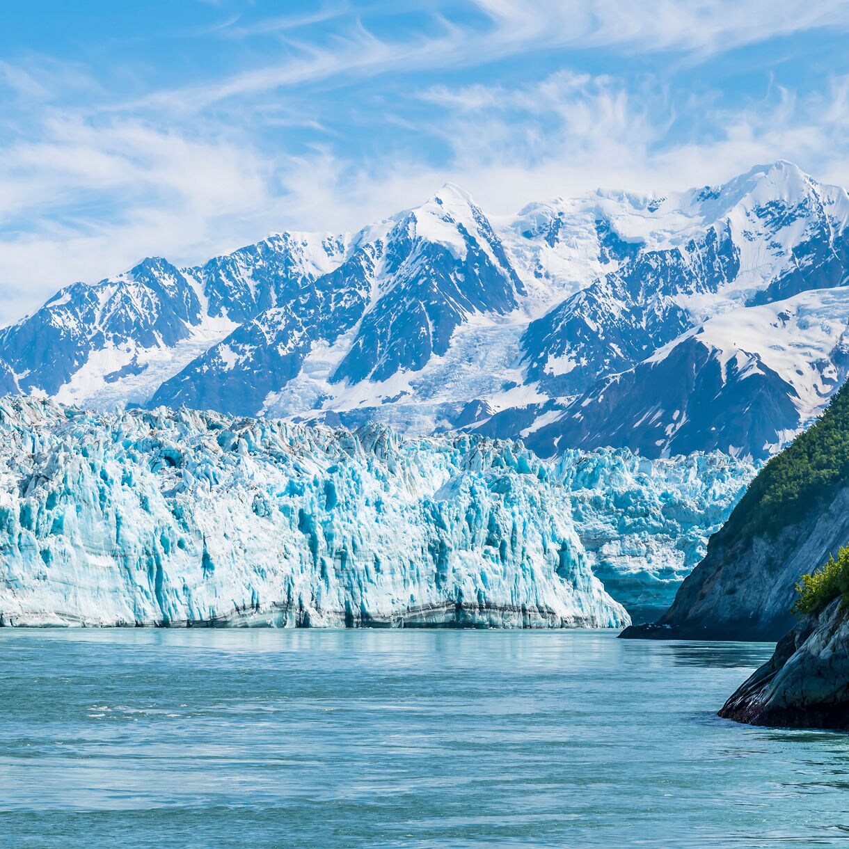 Close-up view of Hubbard Glacier’s towering blue ice wall meeting icy waters, with rocky shoreline in the foreground and rugged snow-streaked mountains in the distance.