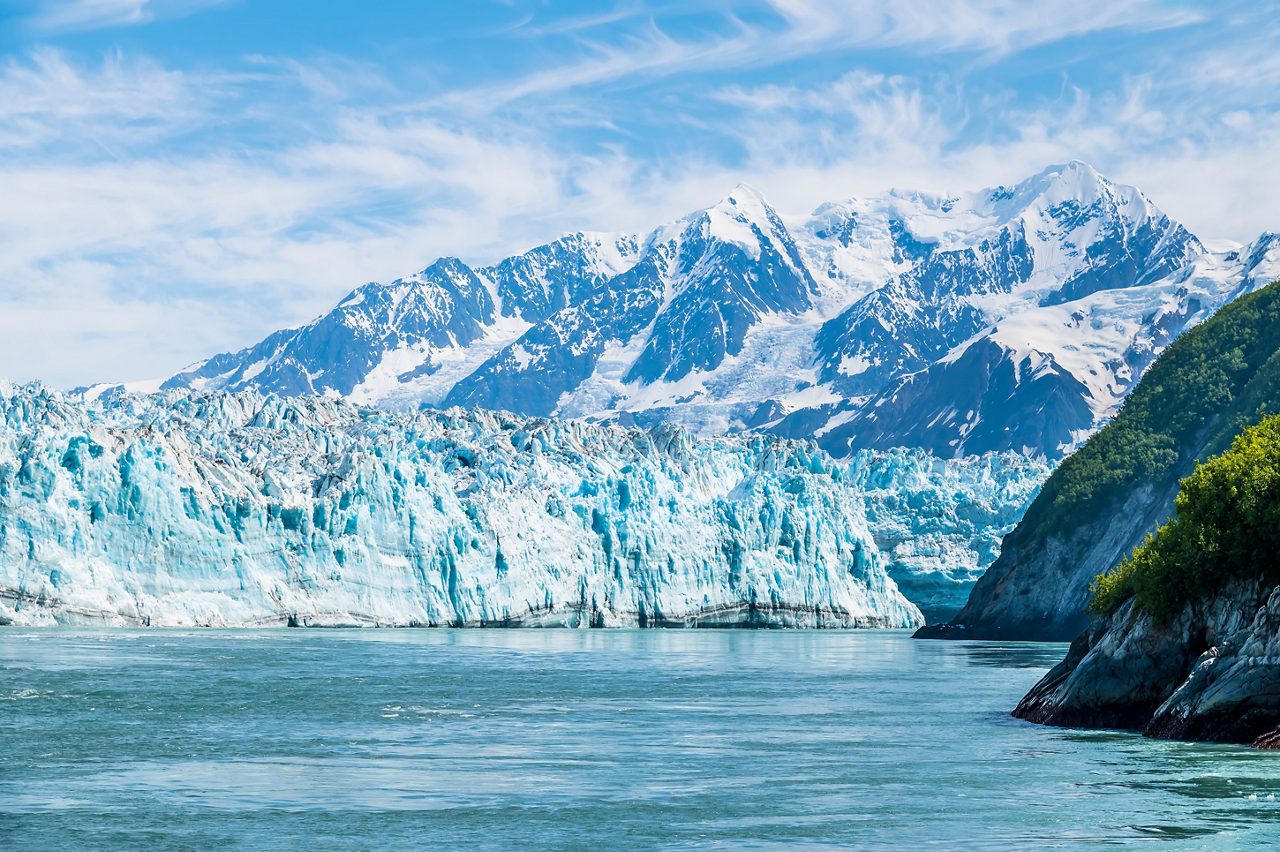 Expansive view of Hubbard Glacier in Alaska with vibrant blue ice fronting a calm bay, flanked by green cliffs and snow-covered mountains under a bright blue sky.
