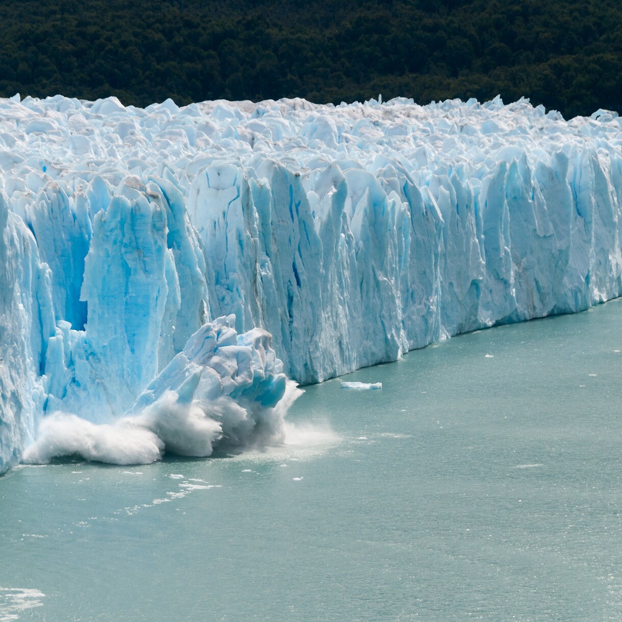 A large chunk of ice calving off the towering face of Hubbard Glacier in Alaska, with snow and ice crashing into the water below against a pale overcast sky.