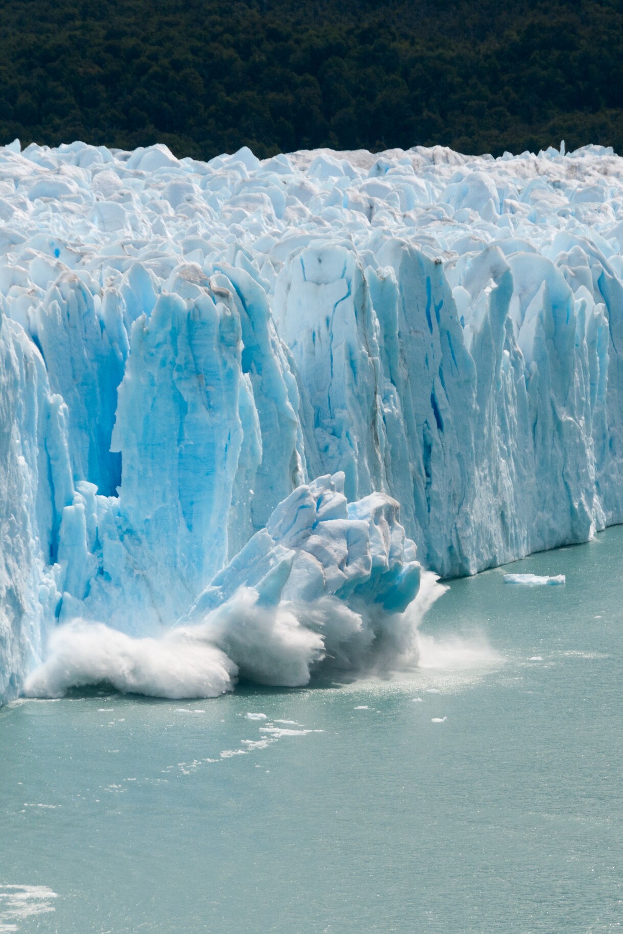 A large chunk of ice calving off the towering face of Hubbard Glacier in Alaska, with snow and ice crashing into the water below against a pale overcast sky.
