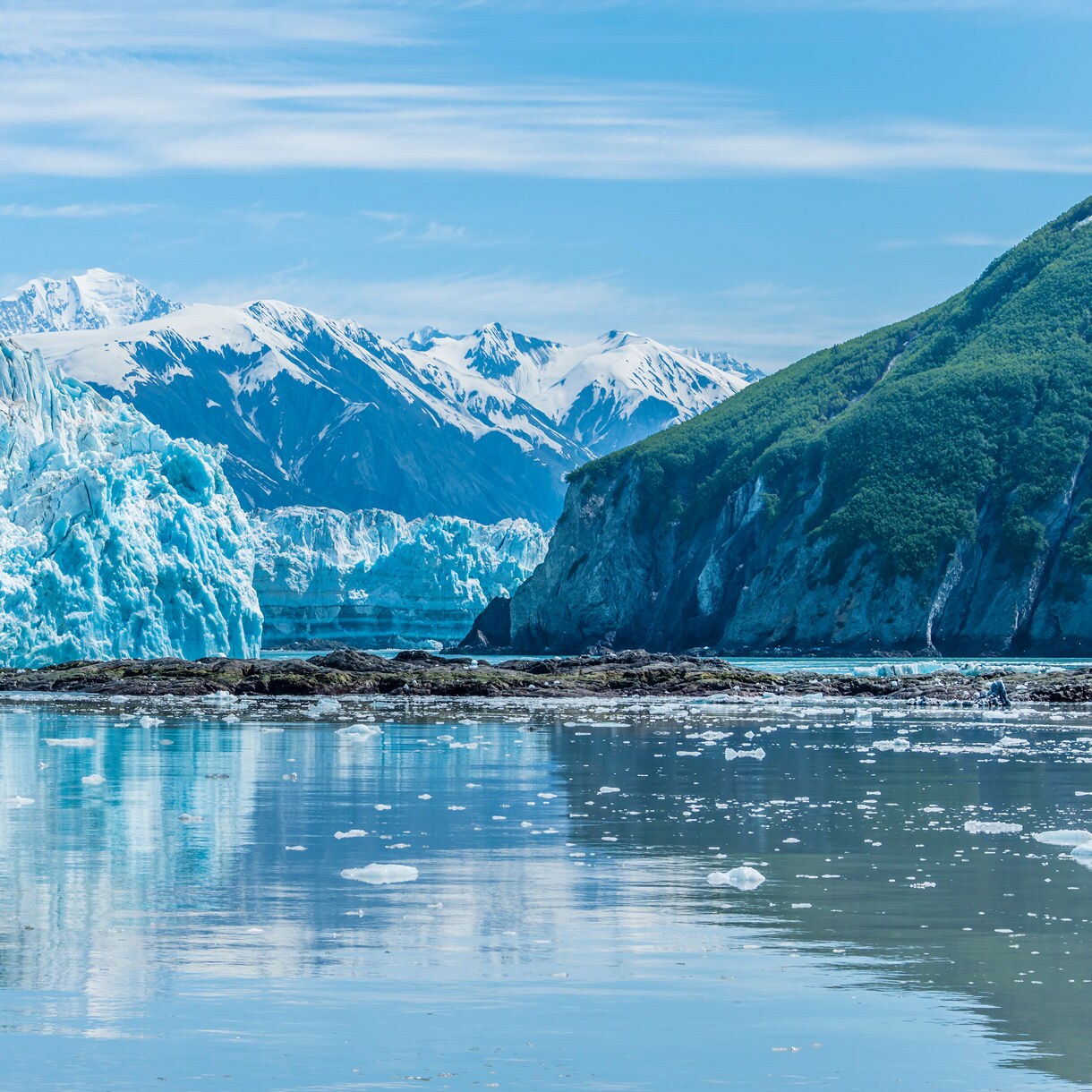 A massive blue glacier wall rising from calm water with snow-covered mountains in the background, small ice fragments floating on the surface, and perfect reflections visible in the water.