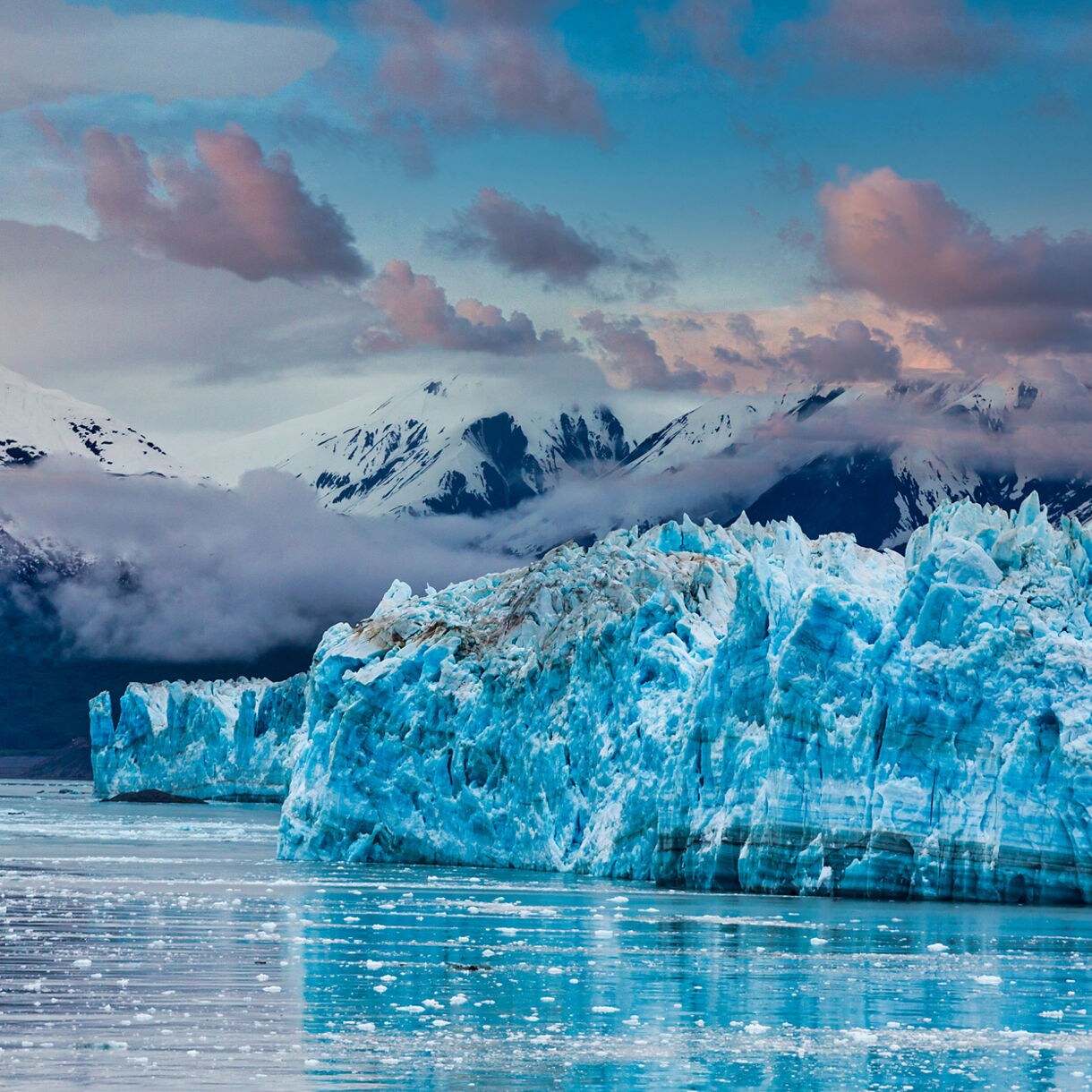 View of Hubbard Glacier in Alaska with its jagged blue ice wall meeting calm waters, backed by snow-covered mountains and a cloudy sky at sunset.