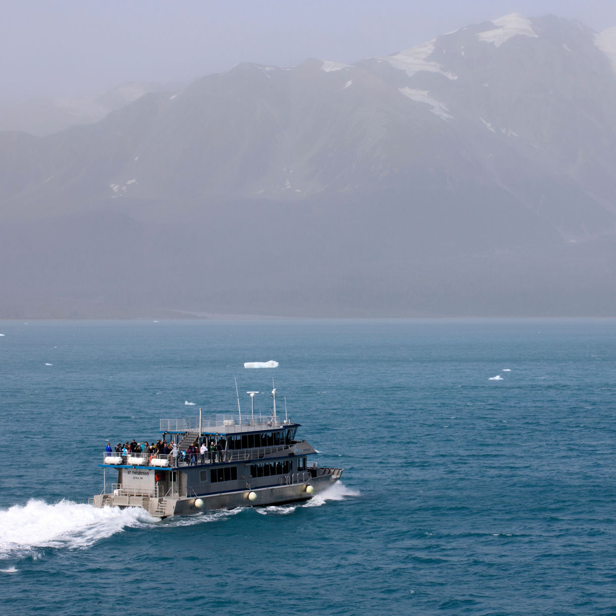 A small tour boat carrying passengers speeds across blue glacial waters near Hubbard Glacier with misty, snow-dusted mountains rising in the background.