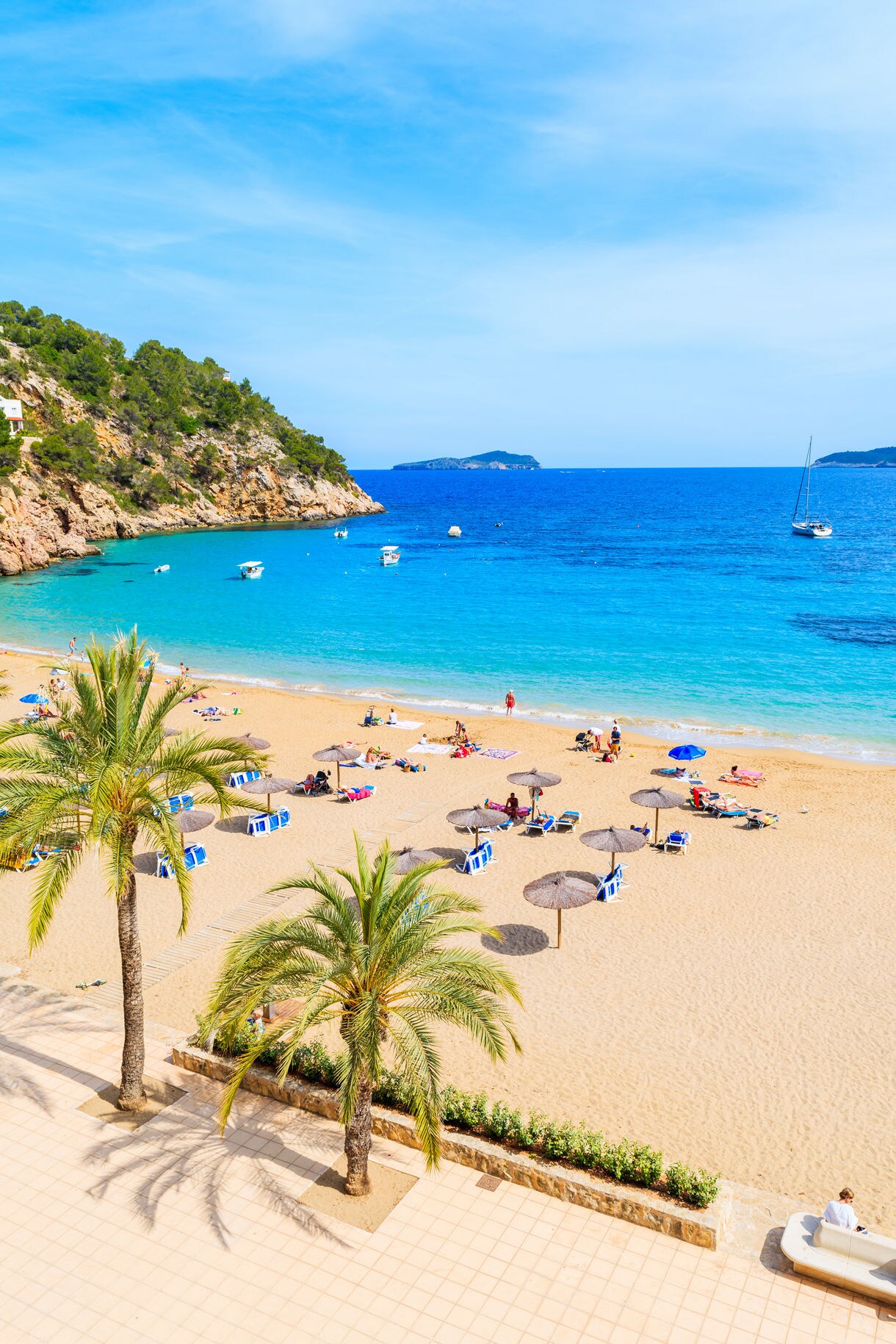 Sunbathers and umbrellas on a sandy beach in Ibiza with palm trees, rocky cliffs, and small boats floating on clear blue water.