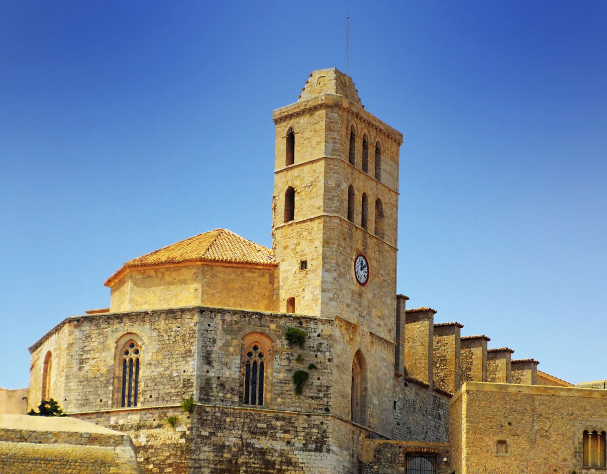 Stone facade of Ibiza Cathedral with a tall bell tower and clock, featuring Gothic and Baroque elements under a clear blue sky.