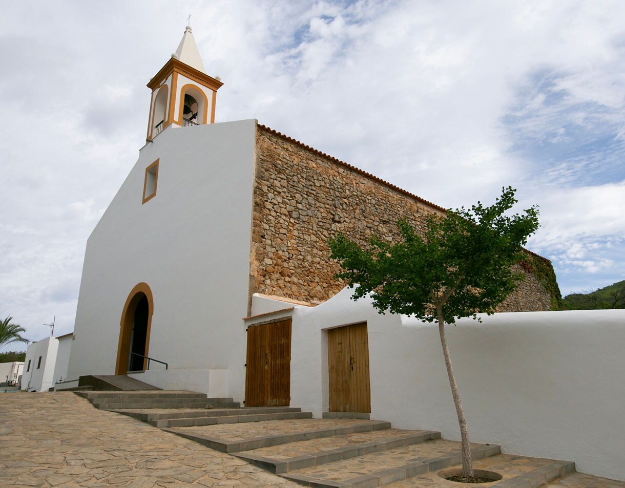 Traditional church in northern Ibiza with whitewashed walls, a stone side wall, and a small bell tower under a partly cloudy sky.