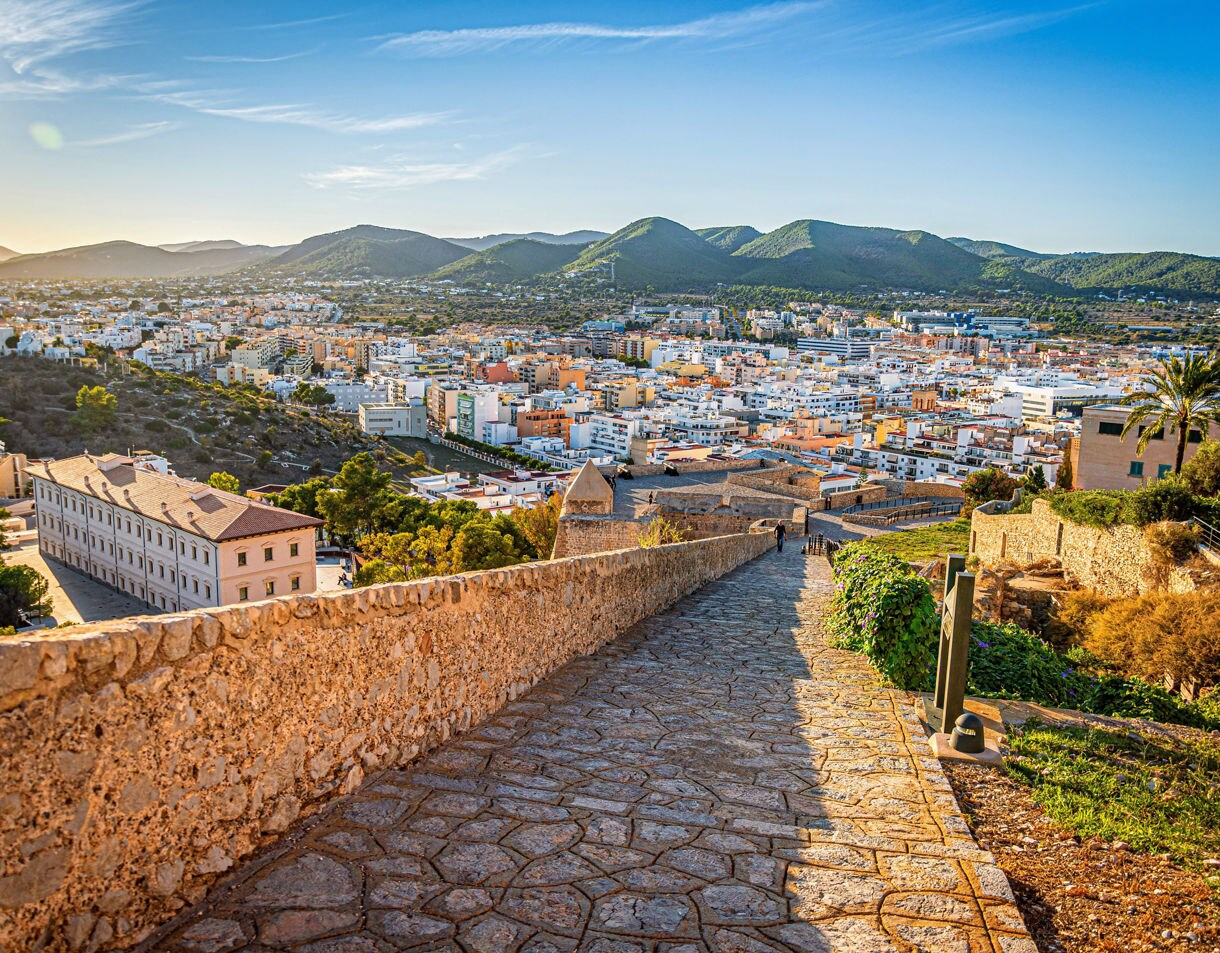 Stone pathway leading down from the historic walls of Ibiza’s Old Town with whitewashed buildings, modern cityscape, and green hills in the distance.
