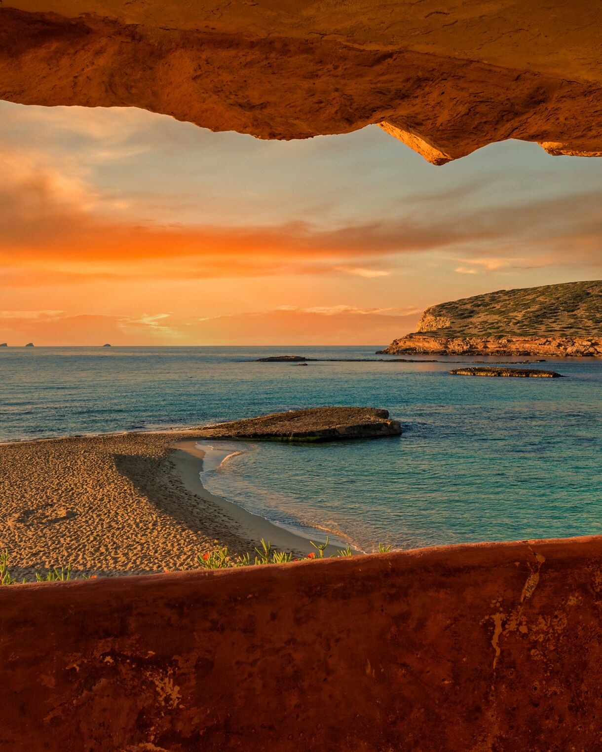 Golden sunset over a quiet Ibiza beach and turquoise sea, viewed through a natural rock opening with cliffs and small islands in the distance.