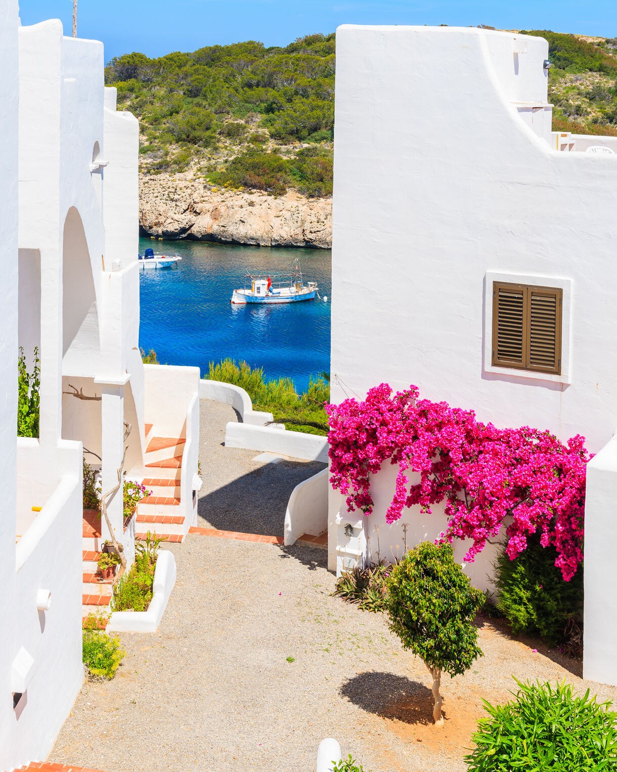 Narrow alley between whitewashed houses in Ibiza with bright pink bougainvillea and fishing boats floating in the blue water below.