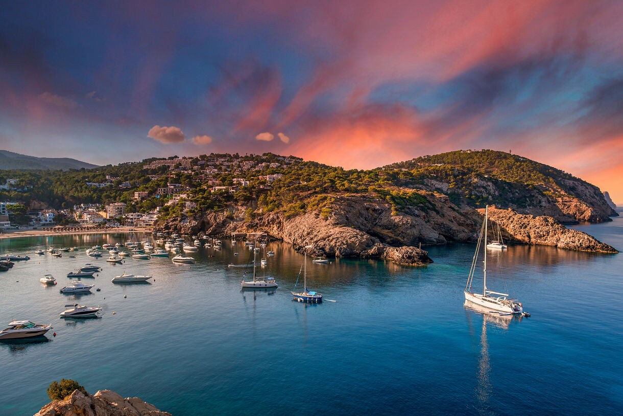 Scenic view of Ibiza’s coastline with rocky hills, scattered villas, and a harbor filled with boats and yachts under a colorful pink and orange sunset.