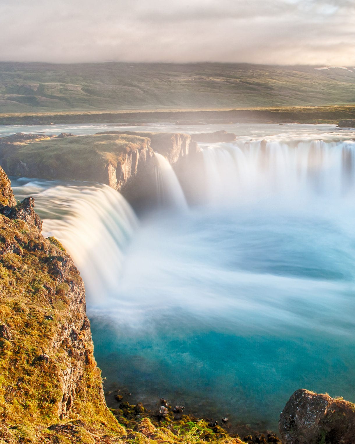 Aerial view of Goðafoss Waterfall in Iceland, with water cascading over a semi-circular cliff into a turquoise pool below, surrounded by rugged terrain and lush greenery.