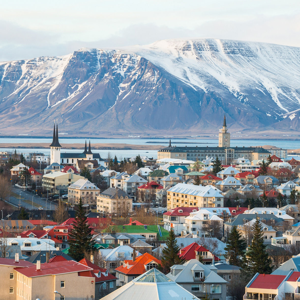 Rooftops and church spires of Reykjavik in front of a bay and a large snow-covered mountain under a clear blue sky.