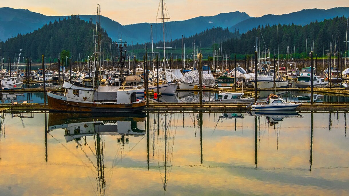 Fishing boats and yachts docked at Hoonah Harbor in Icy Strait, Alaska with colorful reflections on still water, forested hills and mountains in the background at sunset.