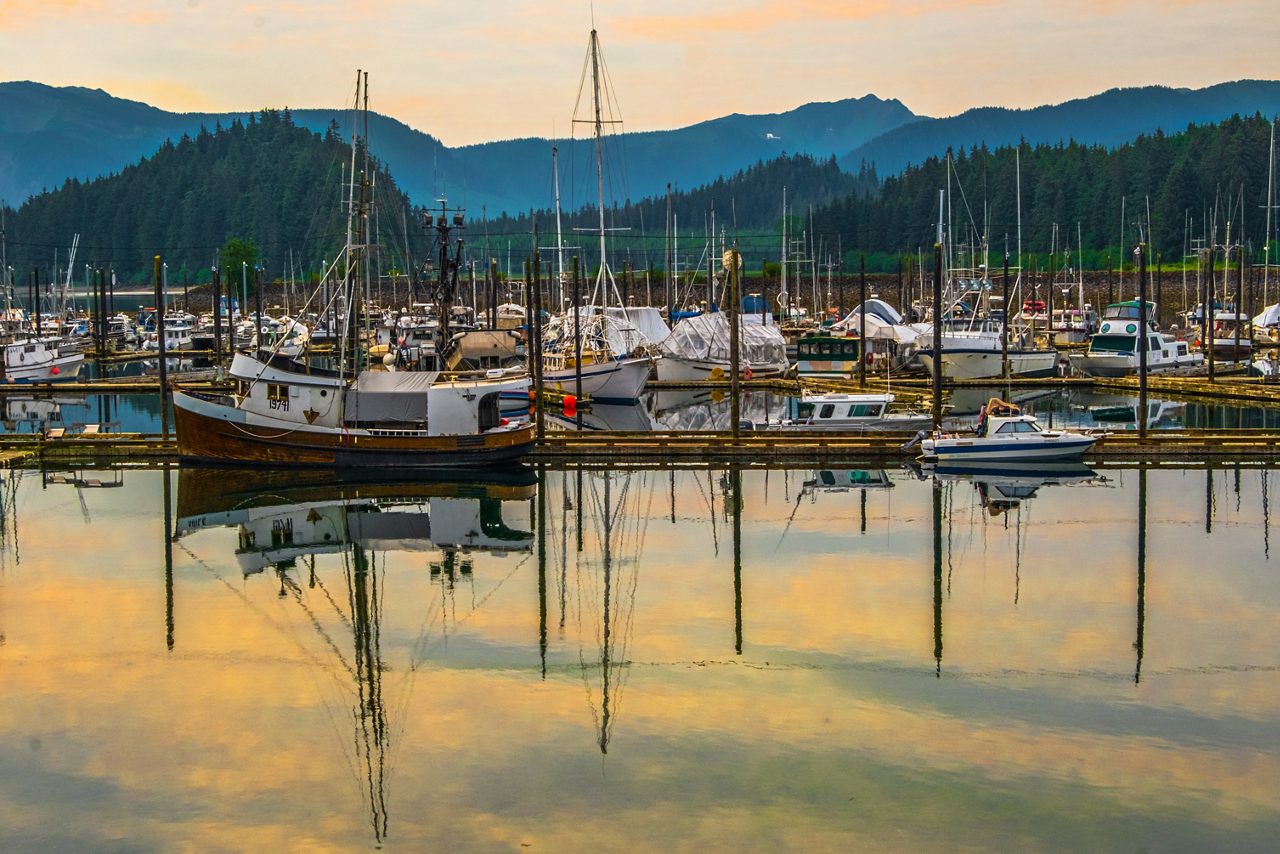 Fishing boats and yachts docked at Hoonah Harbor in Icy Strait, Alaska with colorful reflections on still water, forested hills and mountains in the background at sunset.
