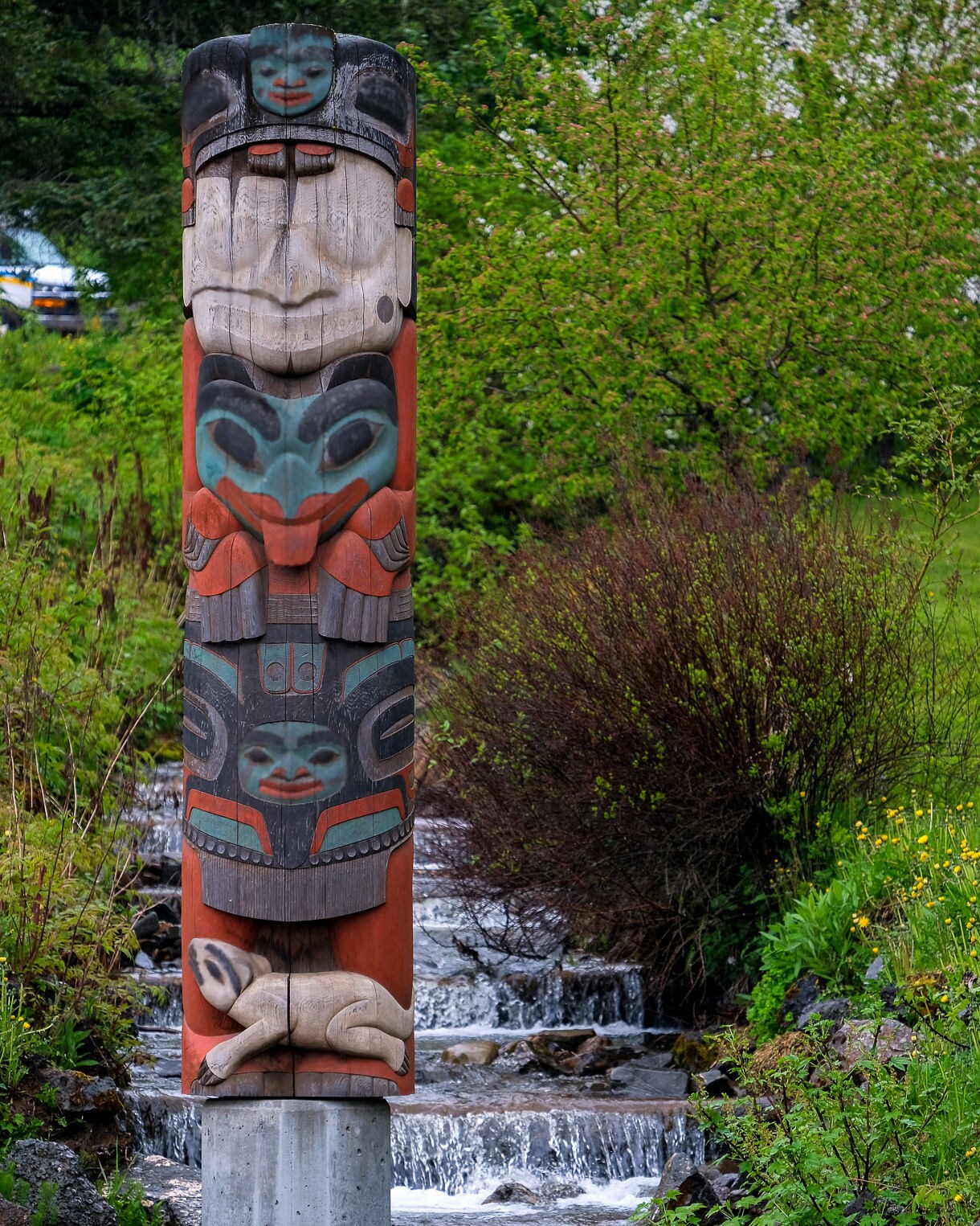 Colorful totem pole carved with human and animal figures set on a concrete base in front of a cascading creek surrounded by lush green foliage in Hoonah, Alaska.