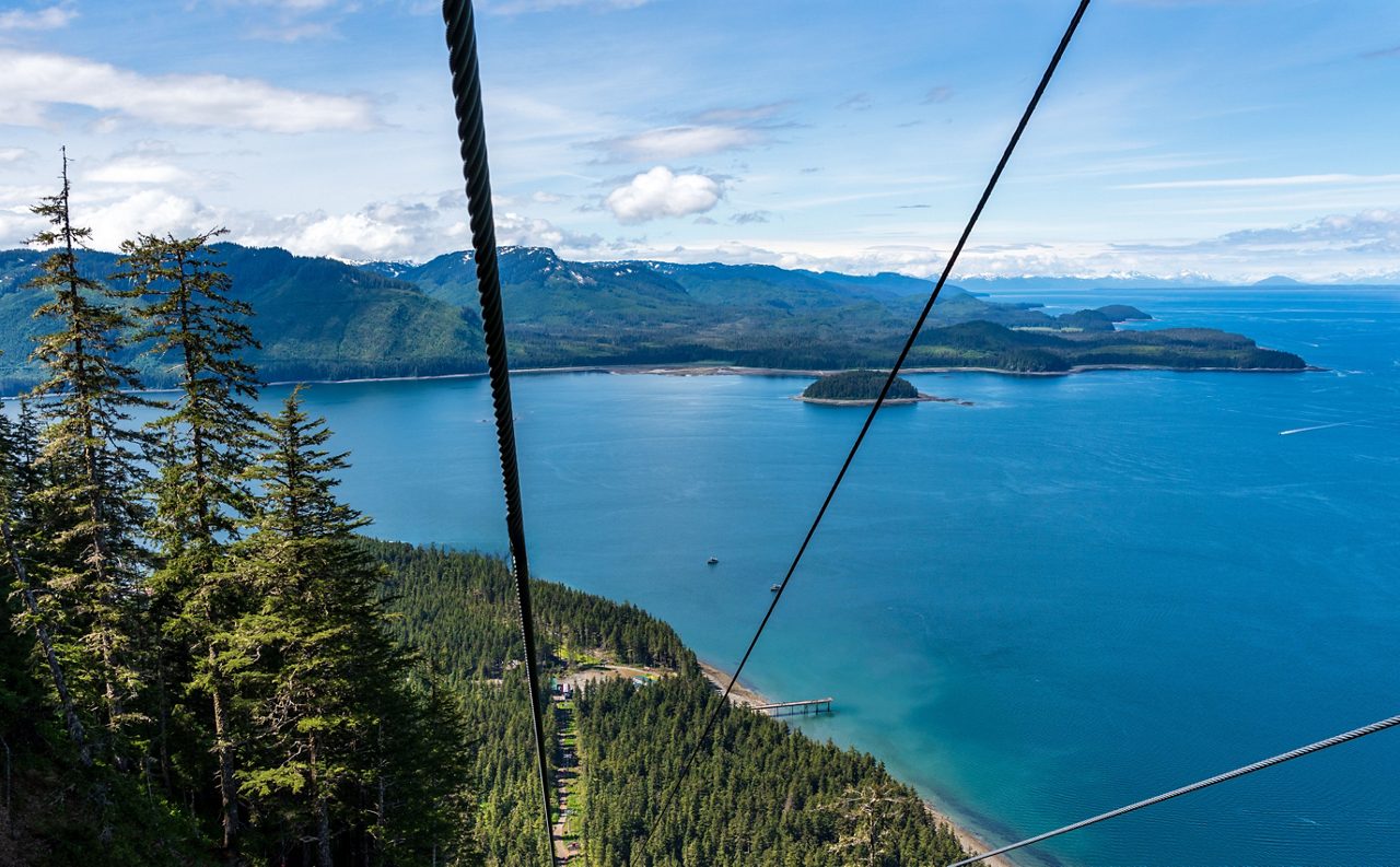 View from the top of Icy Strait Point showing gondola and zipline cables above a forested coastline, with blue ocean, small islands and distant snow-capped mountains.