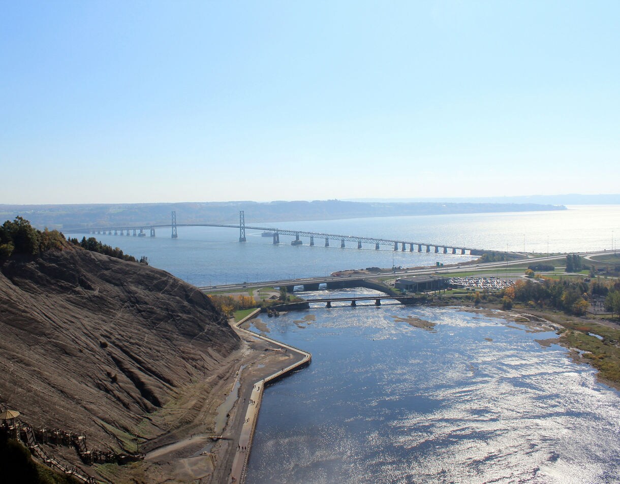 Elevated view of the Île d’Orléans Bridge spanning the St. Lawrence River beside a rocky cliff and shimmering water below.