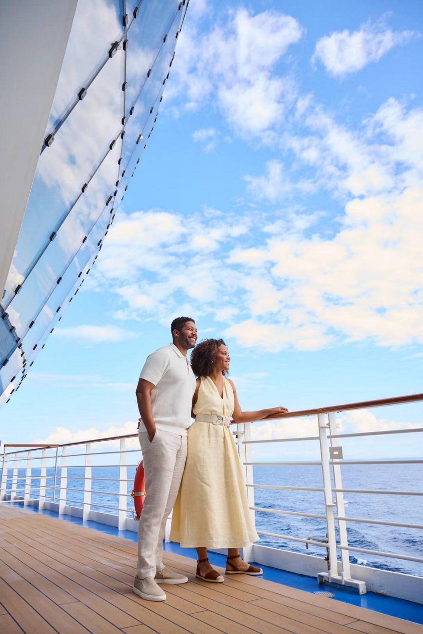 a smiling couple stands at the railing of a Princess® cruise ship, enjoying the open sea and blue sky.