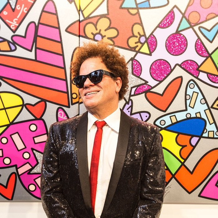 Romero Britto wearing sunglasses, a black sequined jacket, and red tie stands smiling in front of a colorful wall featuring pop art designs with hearts, geometric shapes, and bright patterns. 