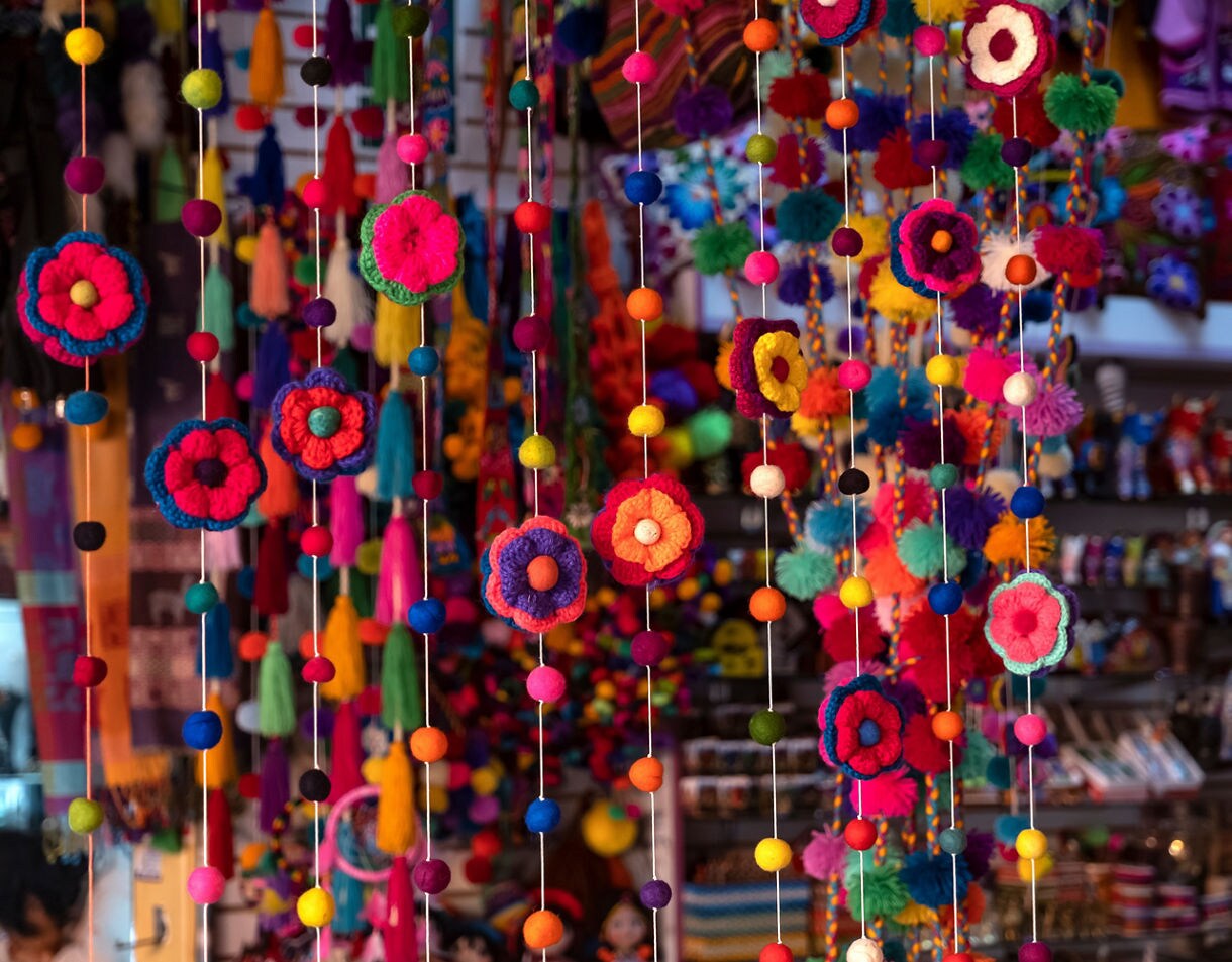 Close-up of colorful yarn flowers and pom-pom garlands hanging in a Peruvian artisan market, with shelves of handmade crafts blurred in the background.