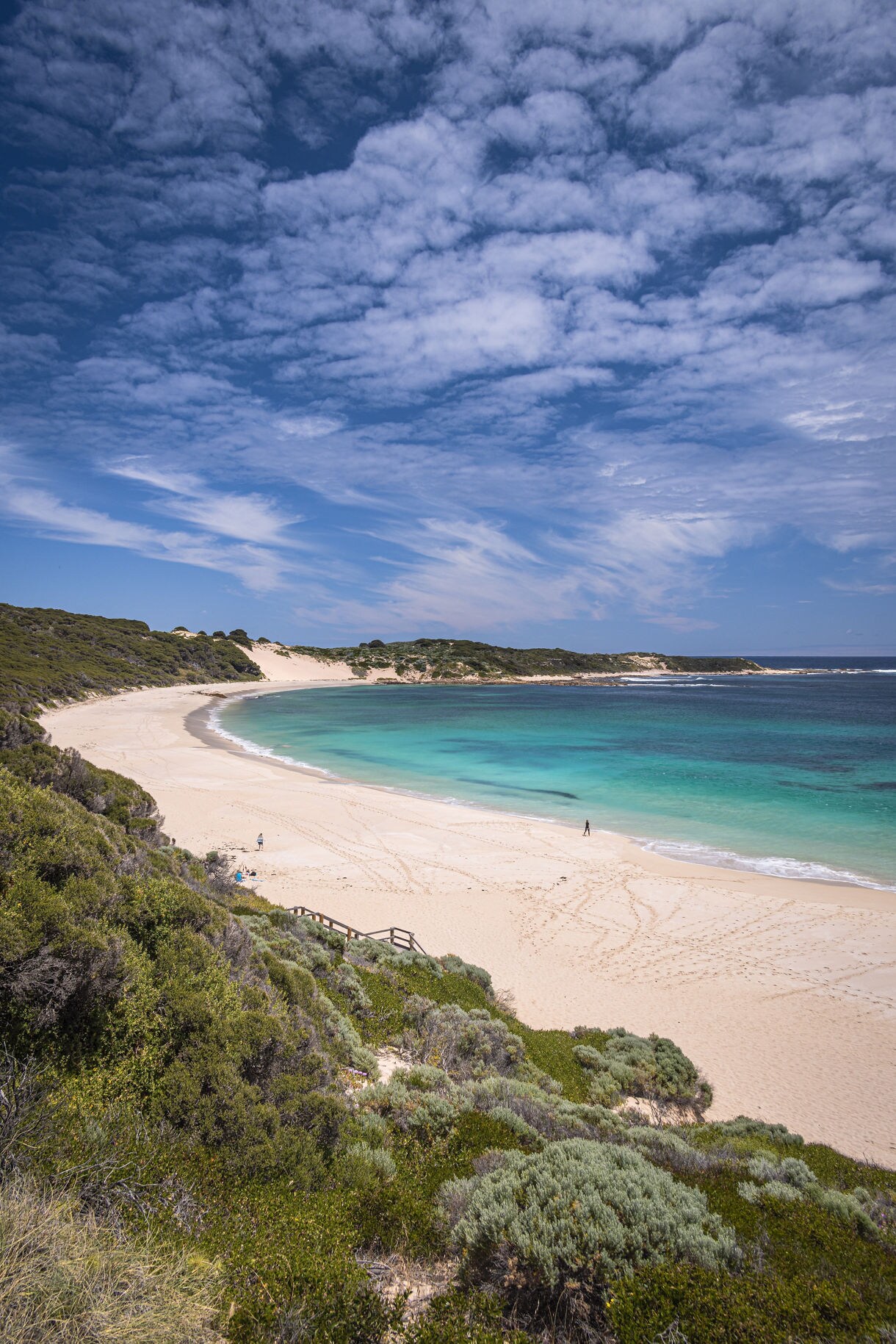 A sweeping white-sand beach with gentle turquoise waves, backed by low green dunes and under a sky filled with scattered clouds.