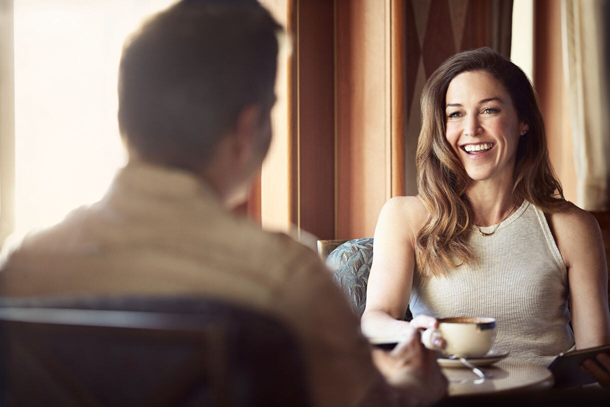 A smiling woman holding a coffee cup seated across from a companion at a warmly lit café table.